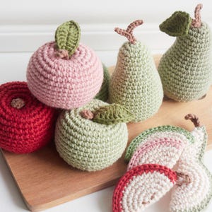 Puede incluir: Juguetes de frutas de crochet en rojo, rosa y verde. Los juguetes tienen forma de manzanas y peras. Hay frutas enteras y rodajas de fruta.
