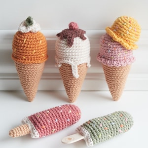 Puede incluir: Conos de helado y paletas de helado de ganchillo en varios colores. Los conos presentan helado naranja, blanco y rosa con adornos decorativos. También se muestran paletas de helado en rosa y verde con palitos de madera. Todos los artículos son hechos a mano.