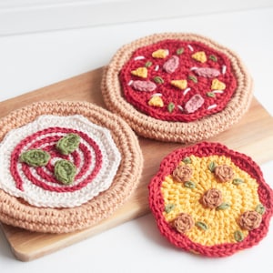 Puede incluir: Tres posavasos de pizza de ganchillo, uno con salsa roja y pimientos verdes, otro con queso amarillo y champiñones marrones y otro con salsa roja y pepperoni rosa.
