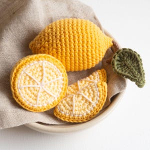 May include: Three crocheted lemons in a bowl. One whole lemon is yellow and the other two are lemon slices with a white center and yellow edges.