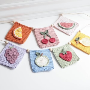 Puede incluir: Una guirnalda de frutas de crochet colorida con un limón, cerezas, uvas, una manzana, una fresa y una sandía. La guirnalda está enhebrada en una cuerda con cuentas de madera.