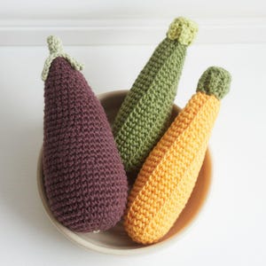 Puede incluir: Tres verduras de ganchillo: una berenjena morada con tallo verde, un calabacín verde y una calabaza amarilla. Están dispuestas en un cuenco marrón claro. Las verduras son hechas a mano y tienen una textura visible.