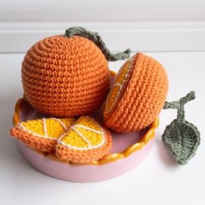 May include: A set of crocheted orange fruit, including a whole orange, a half orange, and three orange slices, displayed on a pink and yellow ceramic dish.
