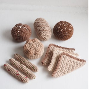 Peut inclure: Un ensemble de jouets en crochet en forme d'aliments, comprenant un pain, un petit pain, un croissant et deux tranches de baguette. Les jouets sont faits dans des tons de brun et beige.