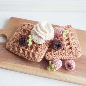 Puede incluir: Una gofre de crochet con un remolino blanco de crochet, acentos de chocolate y fresas rosas sobre una tabla de cortar de madera.