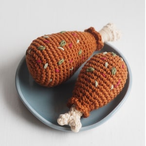May include: Two crocheted chicken legs on a blue plate. The legs are brown with red and green embroidery.