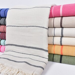May include: A stack of striped towels in various colors, including pink, beige, blue, gray, and green. The towels are folded and stacked on top of each other. The towels have a fringe on the bottom edge.