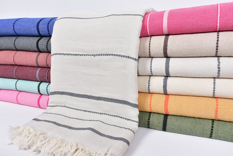 May include: A stack of striped towels in various colors, including pink, beige, blue, gray, and green. The towels are folded and stacked on top of each other. The towels have a fringe on the bottom edge.