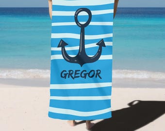 May include: A blue and white striped beach towel with a large black anchor design. The name "GREGOR" is printed in black below the anchor. The towel is held up against a beach and ocean background.