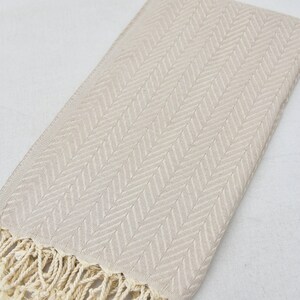 May include: A beige cotton towel with a herringbone pattern and fringe trim.