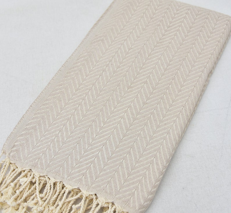 May include: A beige cotton towel with a herringbone pattern and fringe trim.