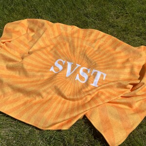 K&ouml;nnte beinhalten: Ein orange-gelbes Strandtuch mit dem Wort "SVST" in wei&szlig;en Blockbuchstaben. Das Handtuch hat ein Sonnenstrahl-Design, das von der Mitte ausgeht. Das Handtuch ist gefaltet und liegt auf gr&uuml;nem Gras.
