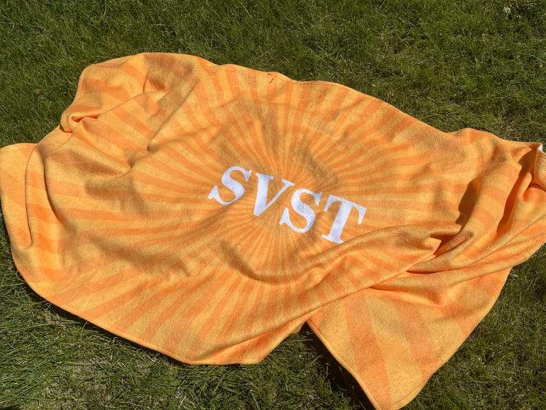 K&ouml;nnte beinhalten: Ein orange-gelbes Strandtuch mit dem Wort "SVST" in wei&szlig;en Blockbuchstaben. Das Handtuch hat ein Sonnenstrahl-Design, das von der Mitte ausgeht. Das Handtuch ist gefaltet und liegt auf gr&uuml;nem Gras.