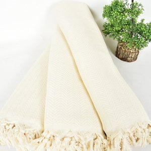 May include: A white cotton throw blanket with a herringbone pattern and fringe trim. The blanket is folded in thirds and is lying on a white surface.