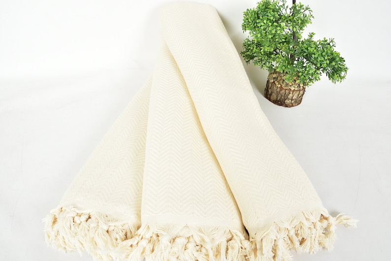 May include: A white cotton throw blanket with a herringbone pattern and fringe trim. The blanket is folded in thirds and is lying on a white surface.