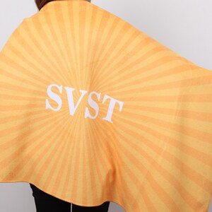 K&ouml;nnte beinhalten: Ein gelb-orange gestreifter Umhang mit den wei&szlig;en Buchstaben "SVST" in der Mitte. Der Umhang ist &uuml;ber die Schultern einer Person drapiert, wobei der Hinterkopf und die Schultern sichtbar sind.
