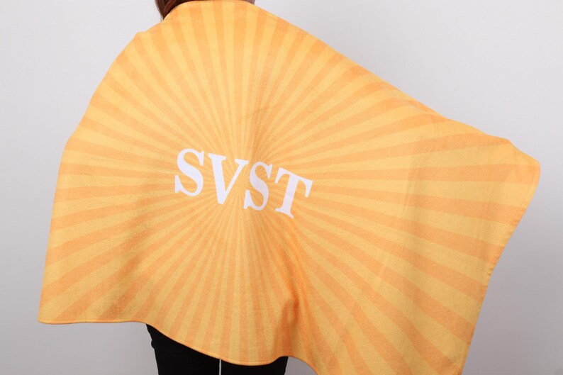 K&ouml;nnte beinhalten: Ein gelb-orange gestreifter Umhang mit den wei&szlig;en Buchstaben "SVST" in der Mitte. Der Umhang ist &uuml;ber die Schultern einer Person drapiert, wobei der Hinterkopf und die Schultern sichtbar sind.