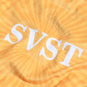 K&ouml;nnte beinhalten: Nahaufnahme eines gelb-orangenen Stoffes mit Sonnenstrahlenmuster. Die wei&szlig;en Buchstaben "SVST" sind deutlich in der Mitte zu sehen. Der Stoff hat ein weiches, strukturiertes Aussehen.