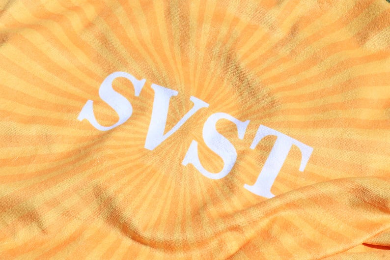 K&ouml;nnte beinhalten: Nahaufnahme eines gelb-orangenen Stoffes mit Sonnenstrahlenmuster. Die wei&szlig;en Buchstaben "SVST" sind deutlich in der Mitte zu sehen. Der Stoff hat ein weiches, strukturiertes Aussehen.