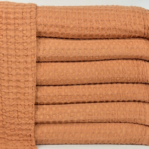 May include: A stack of six waffle weave towels in a warm terracotta colour. The towels have a fringed edge and are made of a soft, absorbent material.