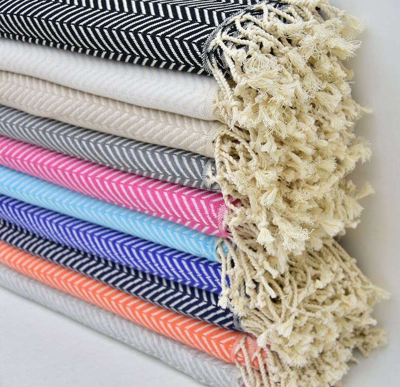 May include: A stack of colorful towels with a chevron pattern and fringe. The towels are folded and stacked in a variety of colors, including black, white, gray, pink, blue, and orange.