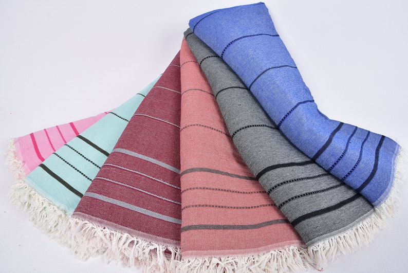 May include: Five striped beach towels in different colors, including pink, light blue, burgundy, coral, and gray. The towels have white fringe around the edges.