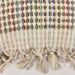 May include: A white Turkish towel with a colorful woven pattern and fringe. The fringe is made of multiple colors of yarn.