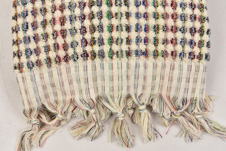 May include: A white Turkish towel with a colorful woven pattern and fringe. The fringe is made of multiple colors of yarn.