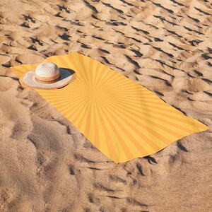 K&ouml;nnte beinhalten: Ein gelbes Strandtuch mit Sonnenstrahl-Design und ein Strohhut auf einem Sandstrand. Das Tuch hat ein strahlenf&ouml;rmiges Muster aus orangefarbenen und gelben Streifen. Der Hut ist beige und braun.