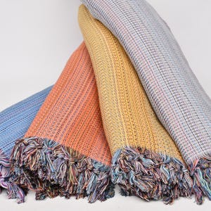 May include: Four woven cotton throws in shades of blue, orange, yellow, and gray. Each throw has a fringed edge with multicolored threads. The throws are folded and stacked, showcasing their textured weave.
