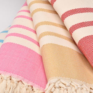 May include: Close-up of several folded, fringed towels in various colours. The towels feature a striped pattern with alternating colours of blue, pink, tan, and red. The towels are made of a woven fabric.
