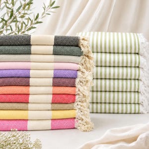 Op de afbeelding: Een stapel rechthoekige handdoeken met een ruitpatroon in zwart, groen, beige, roze, paars, bordeaux, oranje, rood, bruin, geel en magenta, elk met crèmekleurige strepen en franjes, naast een stapel groen gestreepte handdoeken.