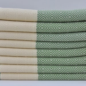 May include: A stack of green and white patterned Turkish towels with fringe. The towels are folded and stacked on a white surface.