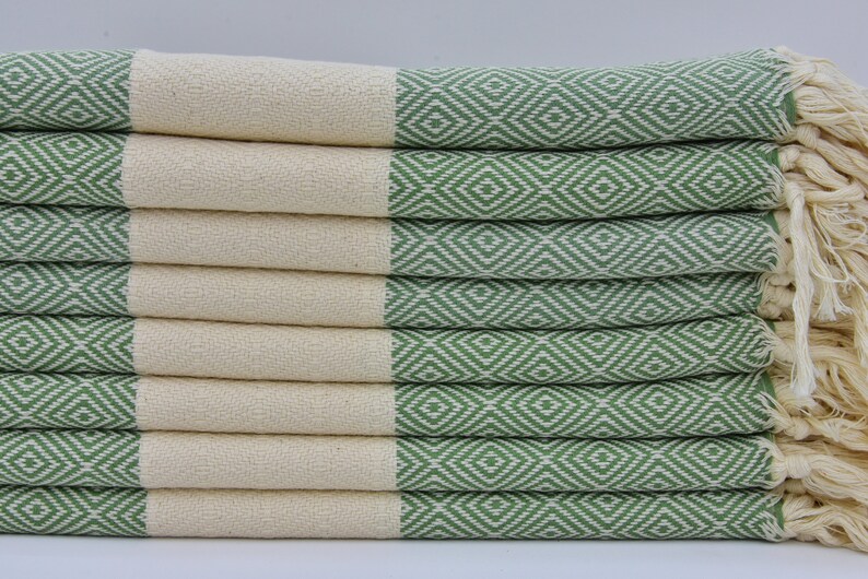 May include: A stack of green and white patterned Turkish towels with fringe. The towels are folded and stacked on a white surface.
