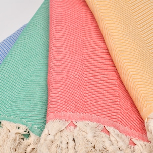 May include: Close-up of four folded beach towels in blue, green, coral, and yellow. Each towel has a chevron pattern and a fringed edge. The towels are made of a soft, woven material.