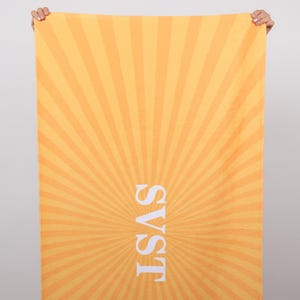 K&ouml;nnte beinhalten: Ein rechteckiges Strandtuch mit einem strahlenf&ouml;rmigen Sonnenaufgangsdesign in Orange- und Gelbt&ouml;nen. Der wei&szlig;e Text "SVST" ist vertikal zentriert. Das Tuch wird von zwei H&auml;nden gehalten.