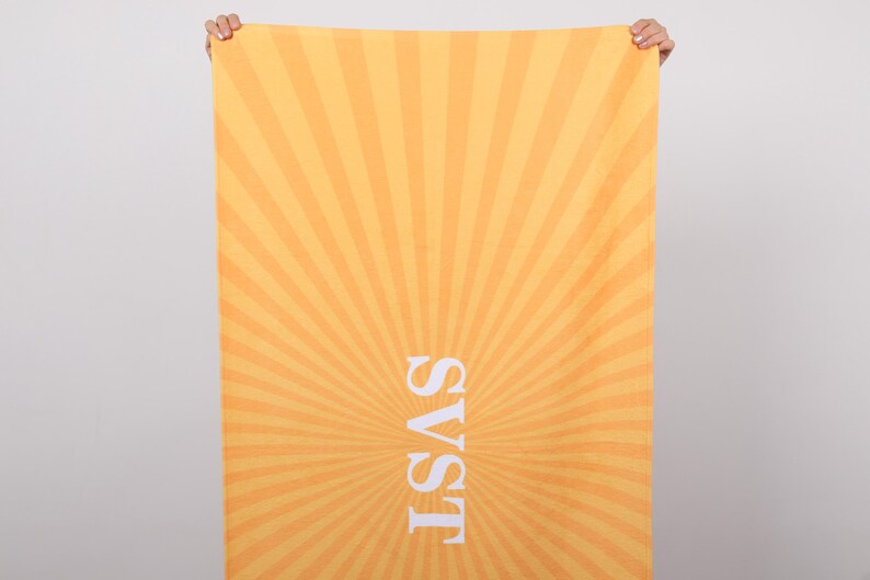 K&ouml;nnte beinhalten: Ein rechteckiges Strandtuch mit einem strahlenf&ouml;rmigen Sonnenaufgangsdesign in Orange- und Gelbt&ouml;nen. Der wei&szlig;e Text "SVST" ist vertikal zentriert. Das Tuch wird von zwei H&auml;nden gehalten.
