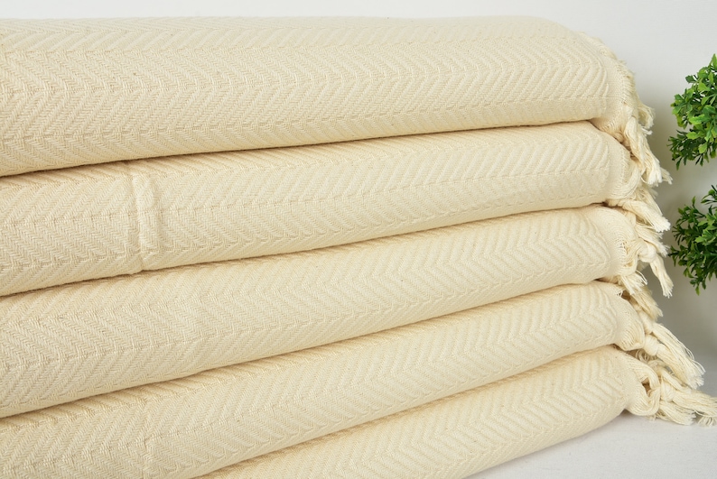 May include: A stack of five cream-colored cotton throw blankets with a herringbone pattern and fringe edges.