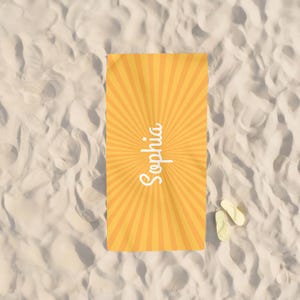 K&ouml;nnte beinhalten: Ein gelb-oranges Strandtuch mit Sonnenstrahlen-Design und dem Namen "Sophia" in wei&szlig;er Schrift. Zwei Paar gelbe Flip-Flops liegen auf sandigem Untergrund neben dem Handtuch.
