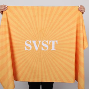 K&ouml;nnte beinhalten: Ein hellorangefarbenes und gelbes Handtuch mit einem Sonnenstrahl-Design. Auf dem Handtuch stehen die wei&szlig;en Buchstaben "SVST" in der Mitte. Das Handtuch wird von einer Person gehalten.