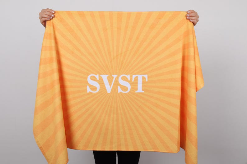 K&ouml;nnte beinhalten: Ein hellorangefarbenes und gelbes Handtuch mit einem Sonnenstrahl-Design. Auf dem Handtuch stehen die wei&szlig;en Buchstaben "SVST" in der Mitte. Das Handtuch wird von einer Person gehalten.