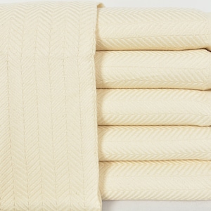 May include: A stack of five cream-colored woven cotton throw blankets with fringe edges. The blankets are folded in a herringbone pattern.