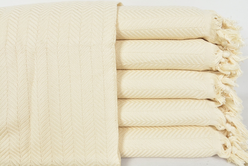 May include: A stack of five cream-colored woven cotton throw blankets with fringe edges. The blankets are folded in a herringbone pattern.