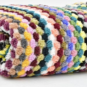 Può includere: Un tessuto multi-rigato e colorato, con una texture a pompon. Il tessuto è piegato a metà e i pompon sono visibili su entrambi i lati.
