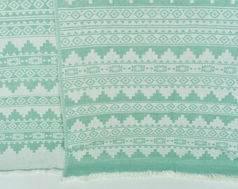 May include: A green and white patterned Turkish towel with fringe on the edges. The towel has a geometric design with white triangles and diamonds on a green background.