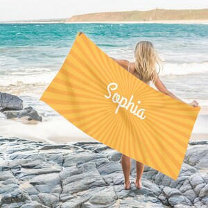 K&ouml;nnte beinhalten: Ein gelb-oranges Strandtuch mit Sonnenstrahlen-Design und dem Namen "Sophia" in wei&szlig;er Schrift. Das Handtuch wird vor einem Strandhintergrund mit Felsen und Meer gezeigt.