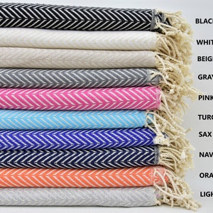 May include: A stack of ten cotton towels with a chevron pattern in various colors, including black, white, beige, gray, pink, turquoise, sax blue, navy blue, orange, and light gray. The towels are folded and stacked with the fringe ends visible.