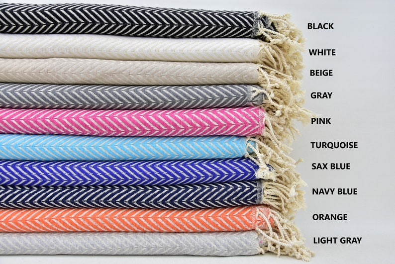 May include: A stack of ten cotton towels with a chevron pattern in various colors, including black, white, beige, gray, pink, turquoise, sax blue, navy blue, orange, and light gray. The towels are folded and stacked with the fringe ends visible.