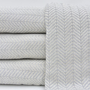 Puede incluir: Una pila de cuatro mantas de lana grises y blancas con estampado de espiga. Las mantas están dobladas y apiladas una encima de la otra.