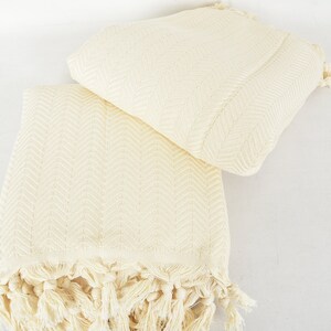 May include: Two cream-colored cotton towels with a herringbone pattern and fringe edges. The towels are folded and stacked on top of each other.